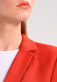 Röd blazer med en strukturerad tygkonstruktion, med fransdetaljer på kragen, parat med en vit ribbad topp under.