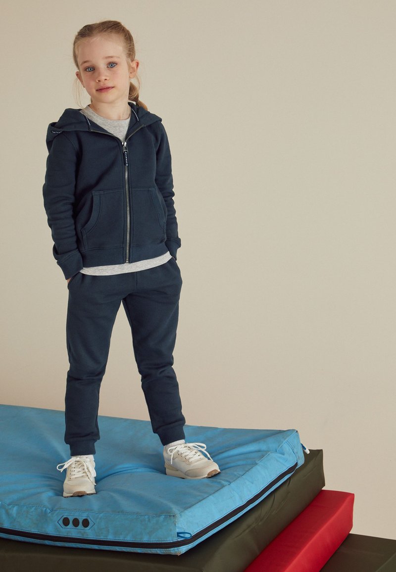 Felpa con cappuccio blu navy con zip e tasche frontali, abbinata a pantaloni della tuta coordinati. Scarpe da ginnastica bianche su un tappetino da palestra blu, con tappetini rossi e verdi sottostanti.