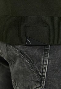 Suéter verde oscuro con dobladillo acanalado, que presenta una etiqueta triangular negra. Vaqueros grises con un detalle de costura distintivo. Apariencia de tejido texturizado.