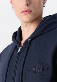Felpa con zip di colore blu navy in pile con una finitura testurizzata, dotata di un logo in rilievo sul petto e di hardware con zip argentato.