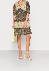 Blumenkleid mit schwarzem Grund und geschichtetem Design. Kurze Puffärmel, V-Ausschnitt und zwei kontrastierende Blumendruckmuster. Hellgrüne Handtasche und Absatzschuhe.