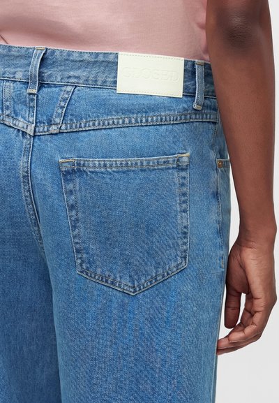 Personne portant un jean bleu clair avec une poche arrière et une étiquette blanche "Closed", chemise rose visible au-dessus de la taille, main sur le côté.