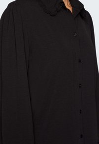 Chemise noire à boutons avec un tissu texturé, col arrondi orné de dentelle, et manches plissées, fermée par des boutons noirs brillants.