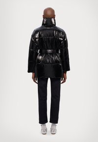 Chaqueta acolchada negra brillante con cuello alto, cintura ceñida y diseño acolchado. Combinada con jeans oscuros y zapatos plateados.