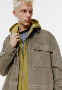 Ruskea ruudullinen villasekoite-paita takki, jossa on nappikiinnitys ja rintatasku, kerrostettuna vihreän hupparin kanssa, jossa on vetoketju ja raidallinen aluspaita.