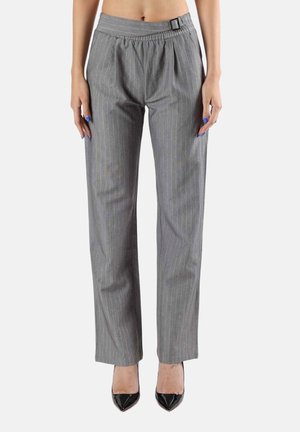 Femme portant un pantalon gris à fines rayures taille haute et jambes droites avec une boucle latérale, associé à des talons noirs à bout pointu, debout sur fond blanc.