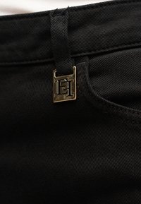 Μαύρο τζιν ύφασμα με λεία υφή, που διαθέτει χρυσό λογότυπο από μεταλλικά στοιχεία και αντίθετη ραφή στη μέση.