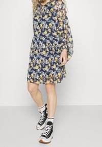 Květinové šaty v námořnické modři s dlouhými průsvitnými rukávy, zdobené žlutými, oranžovými a fialovými květy. Spárované s černými vysokými teniskami.