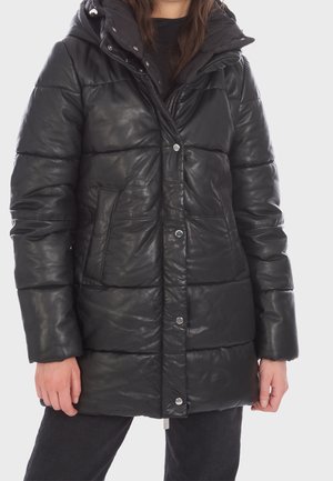 Persona que lleva una chaqueta acolchada negra con botones a presión y cremallera, bolsillos delanteros y un cuello alto sobre un fondo liso.