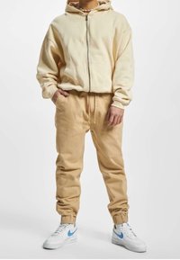 Beige zip-up hoodie met een capuchon, gecombineerd met bijpassende beige joggingbroeken. Witte sneakers met lichtblauwe accenten. Zachte stoftextuur.