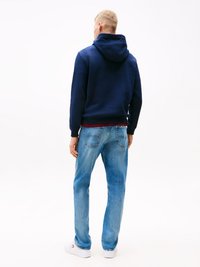 Man wearing a navy blue hoodie, light blue jeans, and white sneakers, standing facing away against a plain white background.