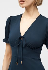 Γυναίκα που φοράει φόρεμα σε σκούρο μπλε χρώμα με κοντά φουσκωτά μανίκια, λαιμόκοψη σε σχήμα V και μπροστινό δέσιμο με κορδόνια με χρυσές άκρες.