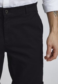 Schwarze Baumwollhose mit glatter Textur, ausgestattet mit einem standardmäßigen Knopfverschluss, sichtbaren Gürtelschlaufen und einer seitlichen Taschenakzentuierung.