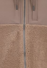 Primer plano de una chaqueta beige que muestra una cremallera metálica frontal con cordones de punto y una tela texturizada tipo lana en la mitad inferior.