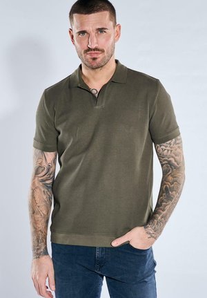 Mann mit kurzen dunklen Haaren und Bart, trägt ein olivgrünes geripptes Poloshirt und blaue Jeans, zeigt Tätowierungen auf beiden Armen.