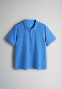 Blauw poloshirt met korte mouwen en een ritskraag, weergegeven tegen een effen lichtgrijze achtergrond.