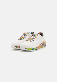 Zapatillas deportivas blancas con acentos en zigzag de colores en la suela y la lengüeta, que presentan cordones multicolores y una superficie texturizada.