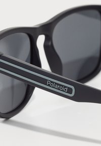 Ochelari de soare negri cu lentile gri tonificate, având un finisaj mat, o formă distinctivă și dungi albastre duble pe lateral cu branding "Polaroid".