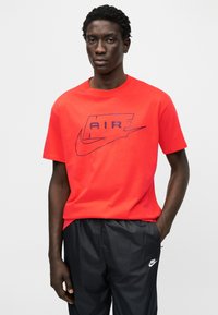 Κόκκινο βαμβακερό μπλουζάκι με το γραφικό "AIR" σε σκούρο μπλε και λογότυπο Nike. Φορεμένο με μαύρο αθλητικό παντελόνι. Σύντομο μανίκι και σχεδιασμός με στρογγυλή λαιμόκοψη.