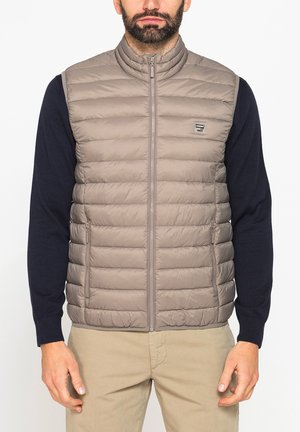 Uomo che indossa un gilet beige trapuntato senza maniche sopra una camicia nera a maniche lunghe e pantaloni beige, in piedi con le mani rilassate lungo i fianchi.