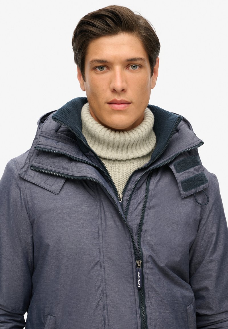 Granatowa kurtka puchowa z zamkiem z przodu, dwoma bocznymi kieszeniami i wysokim kołnierzem, noszona na jasnoszarym swetrze z dzianiny. Podkreślona akcentami z rzepami.