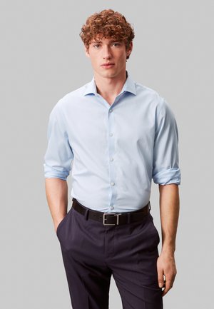 Ung mand med krøllet hår iført en lyseblå skjorte med ærmerne rullede op og mørke bukser, stående mod en ensfarvet grå baggrund.