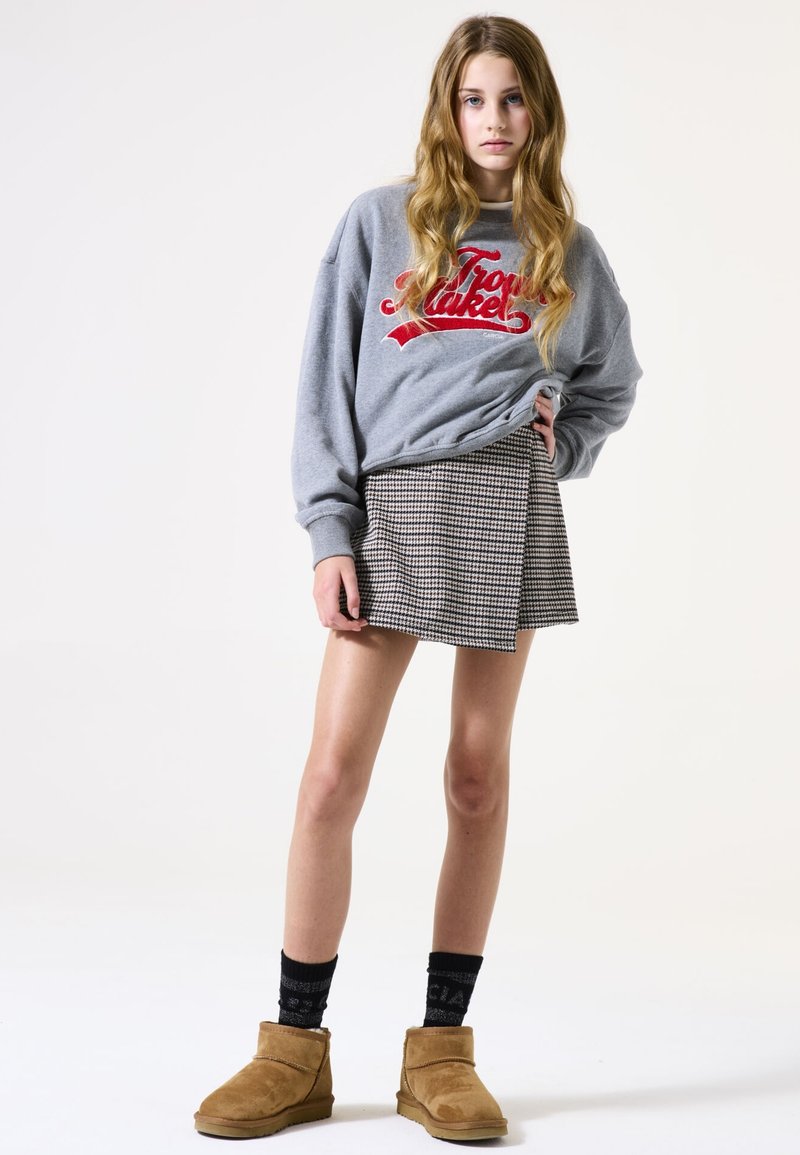 Sweatshirt gris avec texte rouge, jupe portefeuille en pied-de-poule, chaussettes noires à motifs et bottines en suède marron. Modèle posant devant un fond neutre.