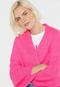 Blonde Frau mit kurzen Haaren, die einen auffälligen, pinken Grobstrickcardigan über einem weißen Shirt trägt und mit verschränkten Armen vor einem weißen Hintergrund posiert.