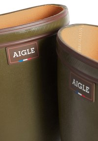 Green rubber boots with brown accents, textured surface, and orange lining. Features branded labels positioned on the outer sides.