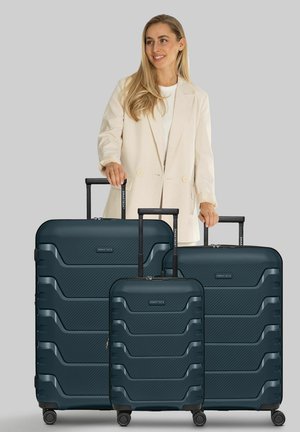 Femme souriante en blazer beige se tient derrière et tient les poignées de trois valises rigides bleu foncé de différentes tailles sur roulettes.