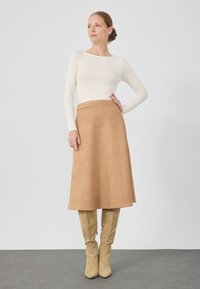 Haut crème à manches longues associé à une jupe en suède beige arrivant au genou et des bottes beiges montantes. Textures lisses, design simple et lignes épurées.