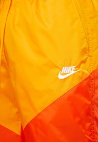 Nike kraťasy v jasně žluté barvě s oranžovým panelem. Vyrobené z lehké látky, na boku mají bílý logotyp.