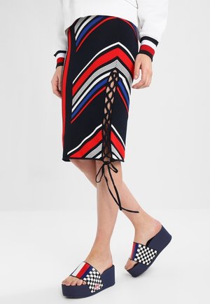 Femme portant une jupe rayée rouge, bleue, blanche et noire avec un détail lacé sur le côté et des sandales compensées à enfiler avec des motifs à carreaux et rayures.