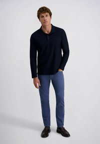 Mann steht da, trägt ein dunkelblaues Langarmpolo, enge blaue Hosen und dunkelbraune Mokassins vor einem schlichten hellen Hintergrund.