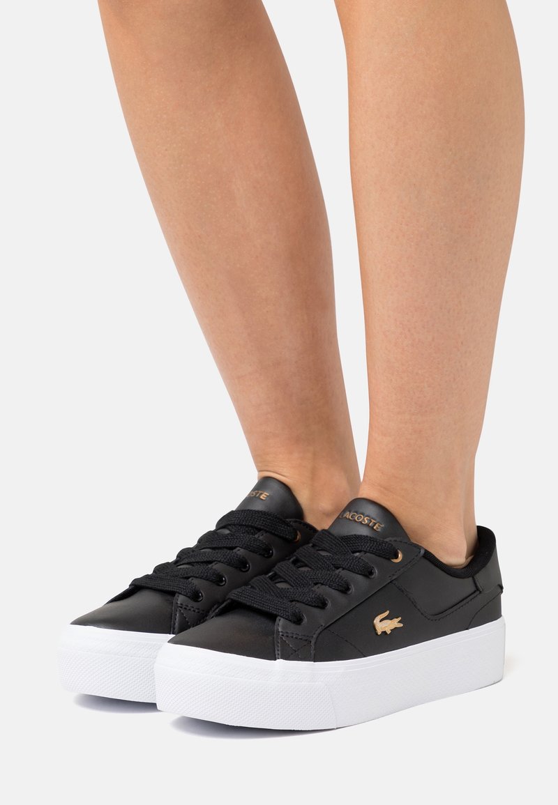 Personne portant des baskets Lacoste noires avec des semelles blanches épaisses, des lacets noirs et un logo crocodile doré sur les côtés, sur un fond clair uni.