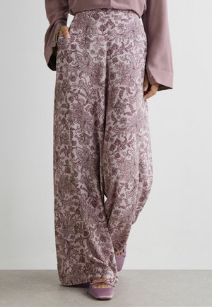 Frau trägt weit geschnittene Lavendelhose mit Blumenmuster, passende lavendelfarbene Ballerinas und eine altrosa Bluse mit langen Ärmeln und ausgestellten Manschetten.