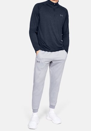 Homme portant une chemise zippée bleu marine à manches longues, un pantalon de jogging gris clair et des baskets blanches, debout les mains dans les poches.