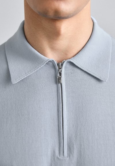 Polo bleu clair à fermeture éclair avec un col côtelé, featuring un détail de fermeture éclair en métal à l'avant. Tissu doux et texturé avec des bords nets.