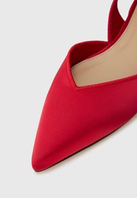 Κόκκινο παπούτσι με μυτερή μύτη και λεία υφή, με κοψίματα στους πλευρικούς τοίχους. Κομψή και λεπτή σιλουέτα, μαύρη σόλα.
