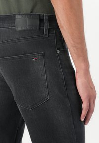 Külevaade inimese puusast, kellel on seljas mustad Tommy Jeans jeans, millel on tagatasku ja bränditud nahklett vöökoht. 