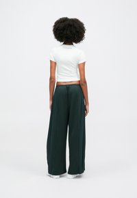 Camisa curta branca de mangas curta, combinada com calças largas e soltas de cor verde escuro com riscas laterais, usadas com sapatos brancos.