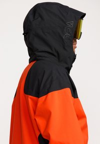 Veste pour homme avec capuche noire et corps orange. Fabriquée en matériau texturé et imperméable, elle arbore un design sportif. Accents zippés remarquables.