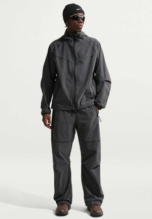 Homme portant une veste et un pantalon de sport noirs, un bonnet noir et des lunettes de soleil, debout les mains détendues, regardant vers la droite.