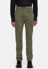Pantaloni chino in cotone verde oliva con vestibilità slim, dotati di tasche frontali e chiusura lampo. Abbinati a scarpe eleganti nere e a una camicia nera con bottoni.