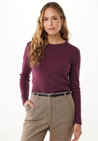 Frau mit langen welligen blonden Haaren, die ein lila Langarmshirt und beigen karierten Hosen mit einem schwarzen Gürtel trägt, steht mit einer Hand in der Tasche.