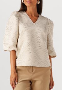Beiger Pullover mit strukturiertem Stoff, V-Ausschnitt, puffierten kurzärmeligen Ärmeln und subtil schimmerndem Muster, kombiniert mit tanfarbenen Hosen.