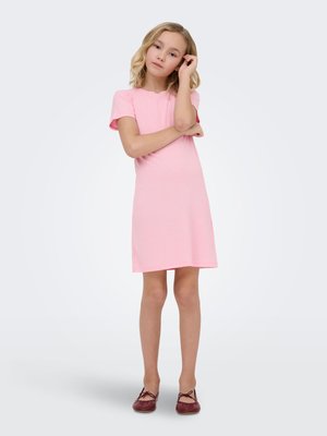 Lyshåret pige iført en lyserød kjole med korte ærmer og bordeaux sko, står med den ene arm over kors og hånden rører hendes ansigt.