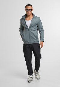 Jasnoniebieska bluza z zamkiem błyskawicznym i wysokim kołnierzem, zestawiona z czarnymi zwężającymi się spodniami. Model ma na sobie sportowe buty i białą koszulkę na ramiączkach.