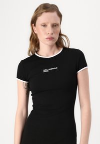 Fekete, rövid ujjú, testhez simuló póló, fehér díszítéssel a nyak- és ujjrészen, amelyen a "KARL LAGERFELD JEANS" logó található a mellkason.