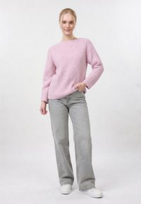 Jeune femme debout, main dans la poche, portant un pull en laine rose, un jean large gris et des baskets blanches, sur un fond uni.